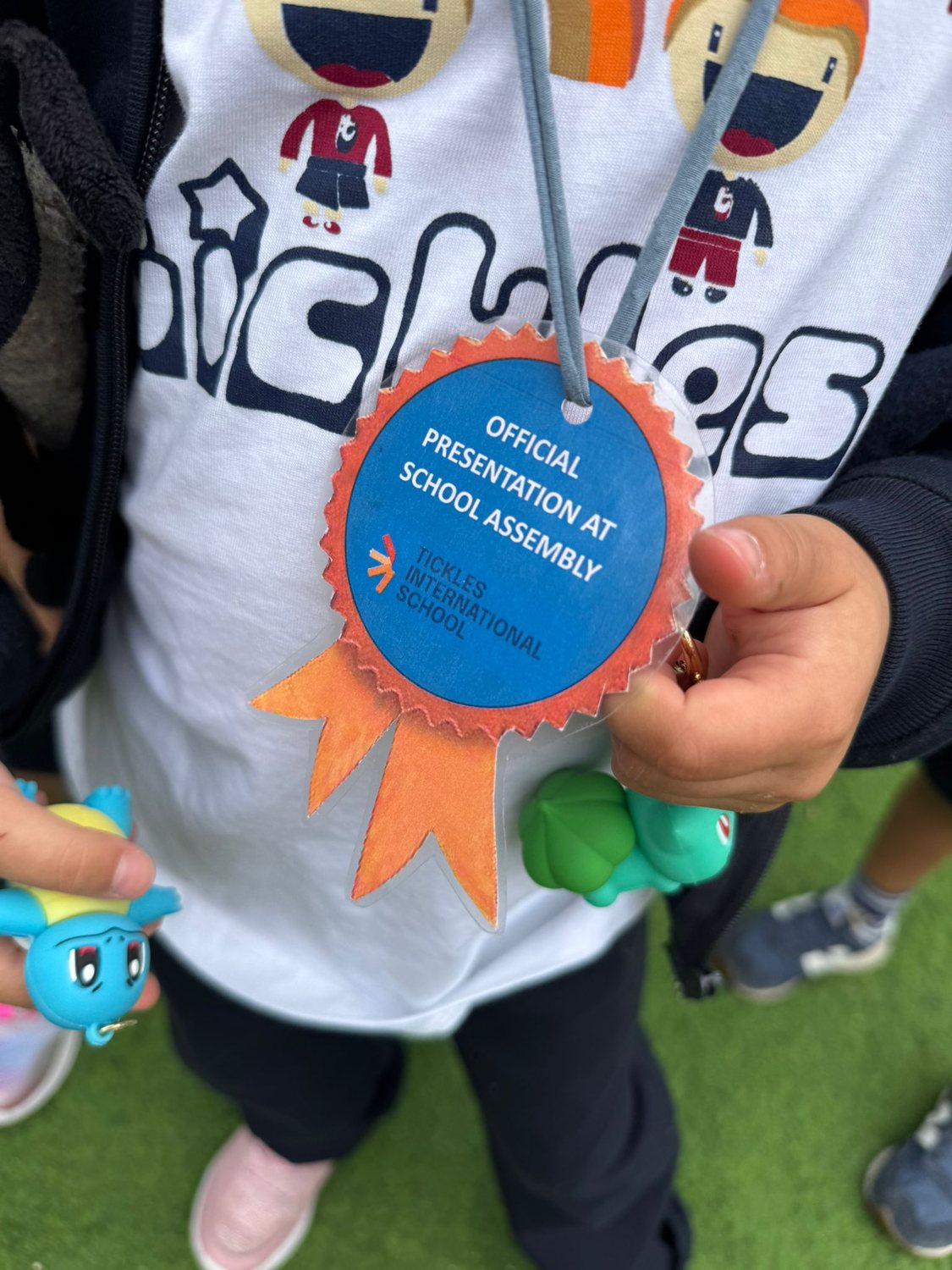 Medalha azul e laranja com a indicação «Official presentation at school assembly», ao pescoço de uma criança com a t-shirt da Tickles
